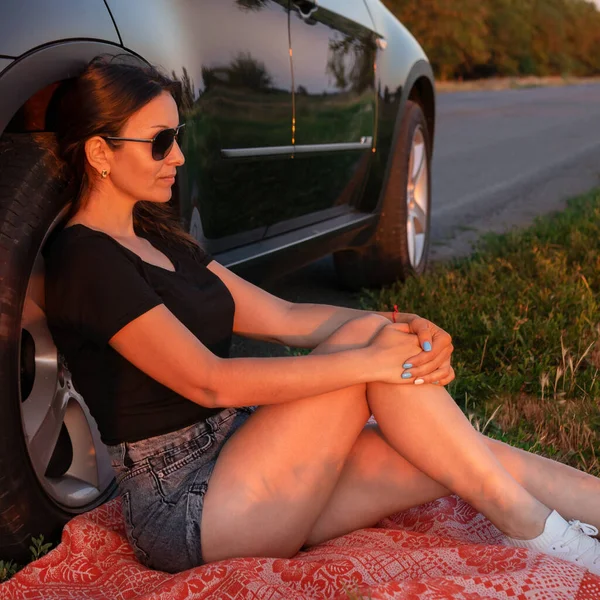 美しい女性が道端の車の近くに座っている 日没を楽しむ官能的な少女 — ストック写真