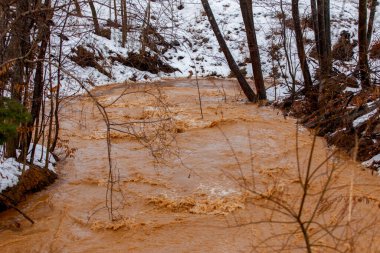 Ekolojik felaket. Kirli, kahverengi bir nehrin akıntısı ağaçların ve karla kaplı toprakların arasında ormanın içinden akar..