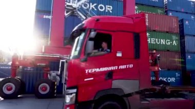Vladivostok, Rusya, 2020 - Limandaki Vladivostok deniz konteynırı terminali operasyonu. Bir kırmızı kamyon limandaki konteynırların yanından geçiyor. Sürücü kabininde güneş parlıyor..