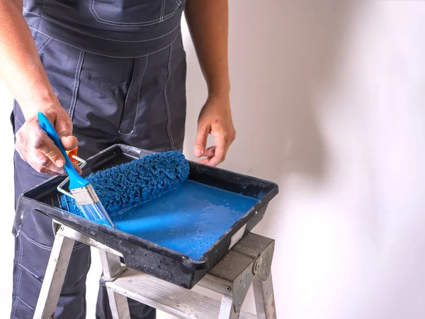 Художник погружает свою кисть в ванну с краской из голубой краски. кювет для роликов на стремянке. Лицензионные Стоковые Изображения