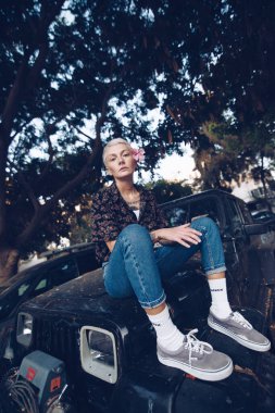 Kot pantolonlu sıradan bir kadın eski siyah bir arabanın üzerinde oturuyor. 