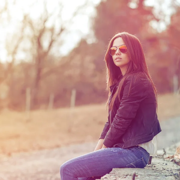 Jeune femme brune sexy en jeans, veste et lunettes de soleil posant — Photo