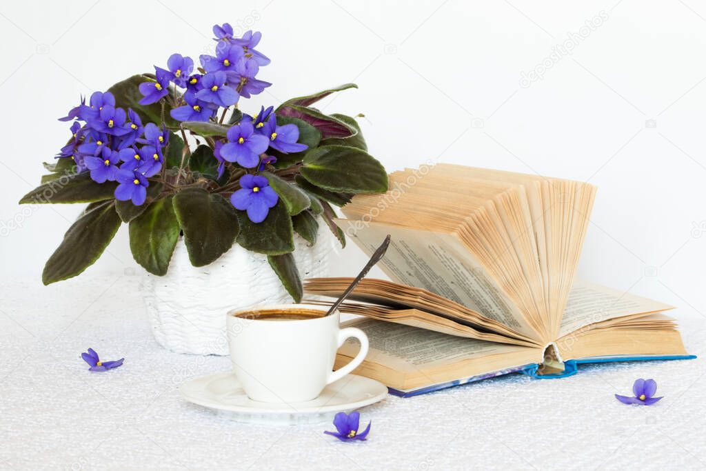 a book with a cup of coffee on a white table and flowers in a basket.
