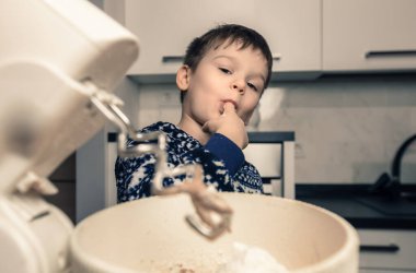 Çocuk mutfakta yemek pişiriyor. Küçük çocuk annesinin yemek hazırlamasına ve elinden hamur ya da krema yalamasına yardım ediyor.