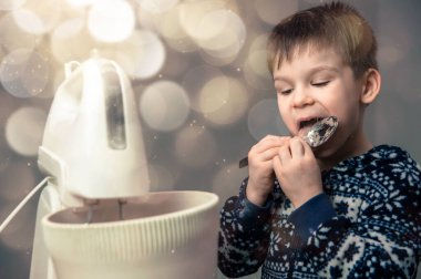 Çocuk mutfakta yemek pişiriyor. Küçük çocuk annesinin yemek hazırlamasına ve kaşıktan hamur veya krema yalamasına yardım ediyor.