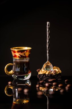 Bir fincan kahve, üzerinde kaşık olan çikolatalı kek ve yansıtıcı siyah yüzey üzerinde kahve çekirdekleri.