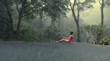 Pembe bikinili güzel bir kadın. Mistik orman manzaralı sonsuz havuzda tropikal yağmurun tadını çıkarıyor.