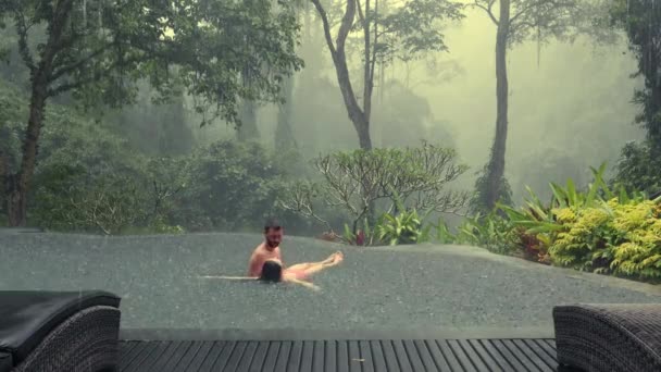 Homem segurando e girando jovem mulher na piscina infiniti com vista para a floresta nebulosa tropical durante a chuva. Belo jovem casal desfrutando juntos na água — Vídeo de Stock