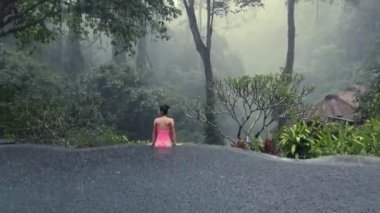 Pembe bikinili güzel bir kadın. Mistik orman manzaralı sonsuz havuzda tropikal yağmurun tadını çıkarıyor.