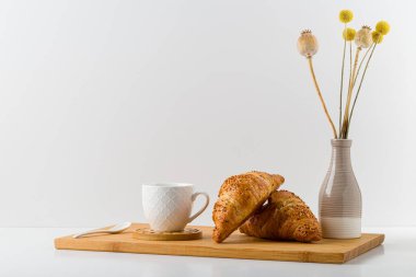 Tepside kruvasan tabakta. Bir fincan sabah kahvaltısı. Vazoda bir buket kurumuş çiçek..