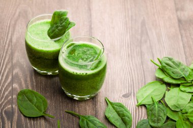 İki temiz bardak ıspanaktan yapılmış yeşil meyve püresi içeriyor. İçkiler ahşap bir masaya konur. Taze ıspanak yaprakları bardakların etrafına saçılmış..