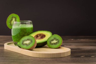 Kivi ve avokadodan yapılan bir içecek ahşap bir tabakta bir bardağa konur. Tam ve dilimlenmiş meyve bardağın yanına dizilmiştir. Arka plan karanlık..