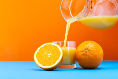 Bir insan cam sürahiden temiz bir bardağa taze portakal suyu döker. İki portakal, camın yanındaki mavi bir yüzeyde dinleniyor. Arkaplan parlak turuncu.