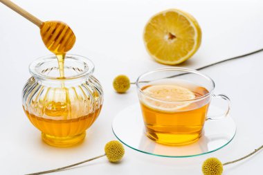 Bir kavanoz bal, limon dilimli bir fincan çayın yanında oturur. Tahta bir kaşık kavanoza bal damlatıyor. Sahnede limonlu ve dekoratif toplar var..