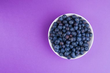 Olgun yaban mersiniyle dolu bir kase mor bir arka planda oturur. Böğürtlenler koyu ve dolgun, abur cubur yemeye ya da tabak ve içeceklere eklemeye hazırlar..