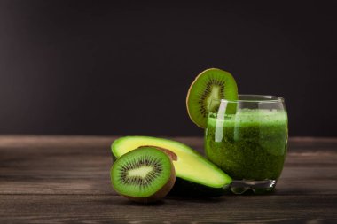 Dilimlenmiş kivi ve ahşap bir masada avokado yanında yeşil içecekle dolu bir bardak vardır. Karanlık arka plan taze malzemelerin altını çiziyor. Bu içecek besleyici ve ferahlatıcı görünüyor..