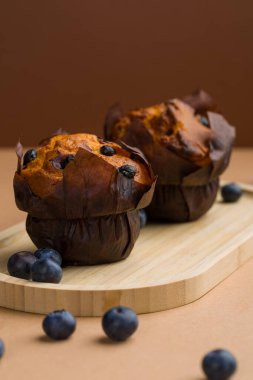 İki yabanmersinli kek, etraflarında dağılmış yabanmersinli ahşap bir tahtanın üzerinde oturuyorlar. Kekler sıcak ve altın kahverengi bir renk sergiliyorlar. Arka plan sade..