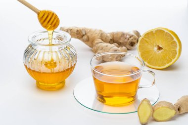 Bir fincan bitki çayı, bal kavanozunun yanında, berrak bir tabakta durur. Taze zencefil ve limon dilimi yakınlarda beyaz bir masanın üzerinde..