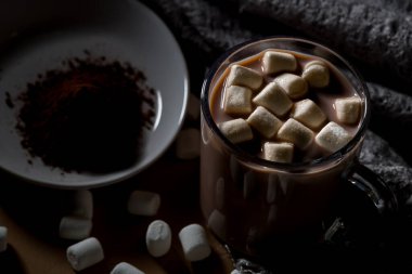 Marşmelovla kaplanmış bir bardak sıcak çikolata ahşap bir masada oturur. Yakınlarda, küçük bir tabakta kakao tozu var. Sahne sıcak bir içkiyi gösteriyor..