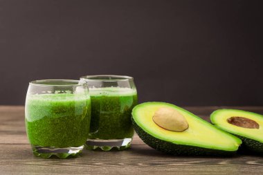 İki bardak yeşil meyve suyu yarım yamalak bir avokadonun yanındaki ahşap masaya konur. Mutfak dekoru ve doğal ışıklandırma mevcut..