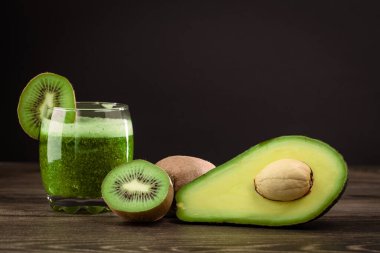 Dilimlenmiş kivi ve yarım avokado 'nun yanında, ahşap bir masanın üzerinde yeşil smoothie ile dolu bir bardak duruyor. Sahnede sağlıklı malzemeler var..