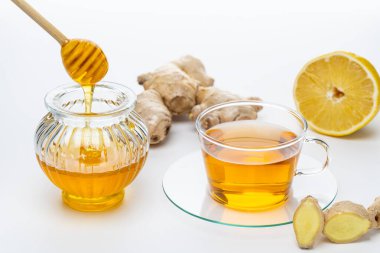 Sıcak zencefil çayı bal ve limonla hazırlanıyor. Bal bir kavanozdan hafifçe ıslanırken cam bardak çayı tutar. Taze zencefil ve limon yarısı yakınlarda..