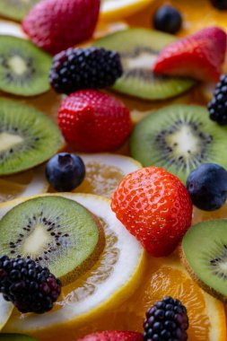 Düz bir yüzeyde çeşitli meyve parçaları düzenlenir. Çilekler, yabanmersinleri ve böğürtlenlerle karışık kivi, portakal ve limon dilimleri renkli bir görüntü oluşturur..