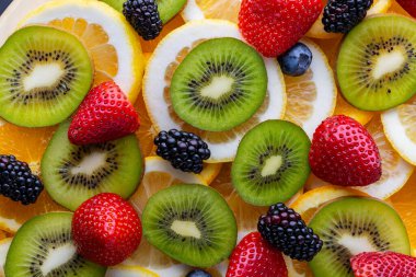 Düz bir yüzeyde kivi, limon ve taze çilek dilimleri böğürtlen ve diğer meyvelerle karıştırılır. Parlak renkler çeşitli meyveleri birlikte gösterir.