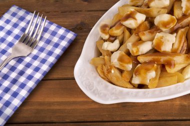 Lezzetli poutine, beyaz bir tabakta, altın patates kızartması, peynir püresi ve zengin kahverengi sosla servis ediliyor. Kırsal ahşap bir masa üzerinde, çatal ve mavi kareli peçeteyle. Düz konsept yemek..