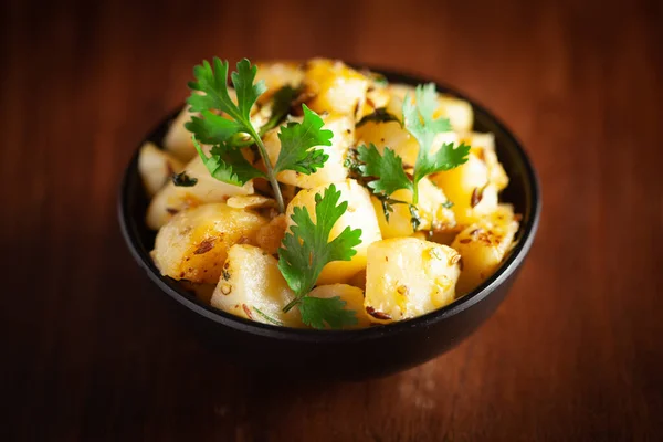 Close-up of Indian vegetarian classic dish Jeera Aloo - Potatoes Flavored with Cumin  garnished with green coriander fresh leaves. Served in black ceramic bowl over wooden background