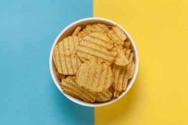 Kırışık Patates cipslerini beyaz seramik kaseye koy, Popüler, gevrek, tuzlu, soluk-sarı renkleri mavi arka planda yemeye hazır