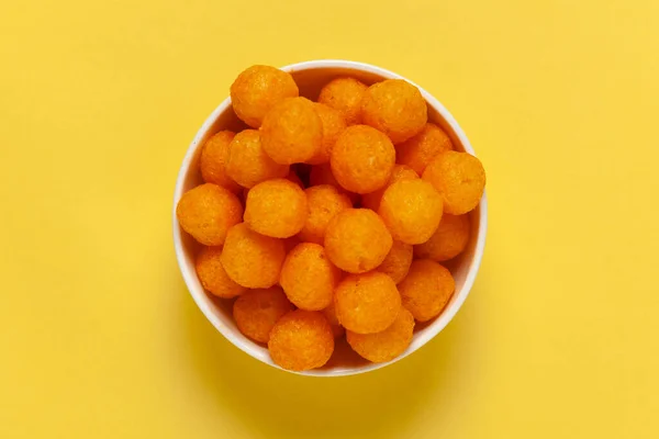 Close up of Cheese Potato Puff Ball Snacks, tangy orange color, Popular Ready to eat crunchy and puffed snacks,  salty in white ceramic bowl over yellow background
