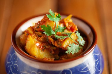 Yeşil kişniş yapraklarıyla süslenmiş baharatlı patates ve domatesli Hint vejetaryen yemeği. Ahşap arka planda seramik handi olarak servis ediliyor..