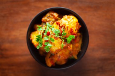 Yeşil kişniş yapraklarıyla süslenmiş baharatlı patates ve domatesli Hint vejetaryen yemeği. Ahşap arka planda siyah bir seramik kasede servis edilmiş..