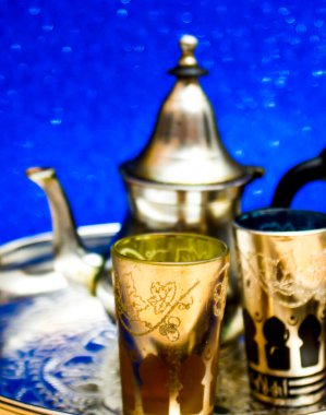 Bir grup çaydanlık ve bir bardak oryantal çay mavi arka plandaki bir tepside parlıyor.