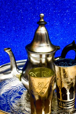 Bir grup çaydanlık ve bir bardak oryantal çay mavi arka plandaki bir tepside parlıyor.