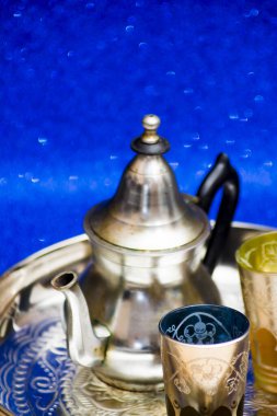 Bir grup çaydanlık ve bir bardak oryantal çay mavi arka plandaki bir tepside parlıyor.