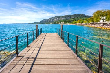 İtalya 'nın Garda köyündeki Garda Gölü' nün mavi sularına uzanan modern bir ahşap iskele manzarası. Arka planda restoranlar olan Lakeside gezinti alanı.