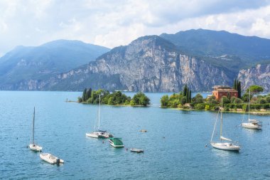 Malcesine yakınlarındaki Val di Sogno körfezinde demirli yelkenli teknelerin manzarası. İtalyan manzarasında Garda Gölü, Veneto 'nun ikonik dağ tepeleri yer alır.