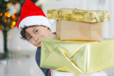 Noel Baba şapkalı kıvırcık saçlı çocuk elinde Noel süsleriyle evde hediyeler tutuyor.