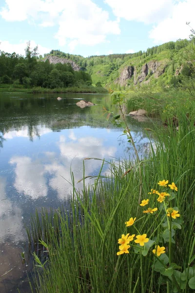 Gölde bulutlu mavi bir gökyüzünün yansıması tepeler ve kayalarla çevrili, yazları sarı çiçekler...