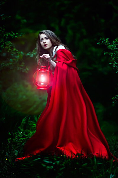 Beautiful woman with red cloak and lantern in the woods by night