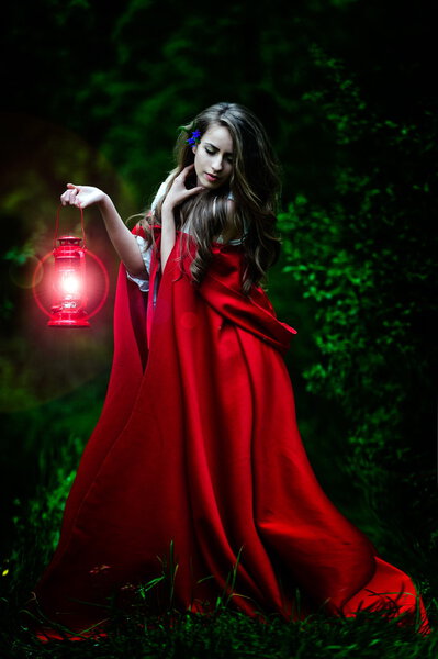 Beautiful woman with red cloak and lantern in the woods by night