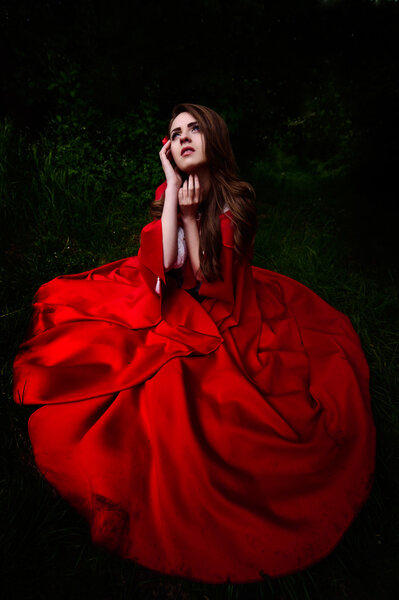 Beautiful woman in red cloak in the woods, sitting on ground