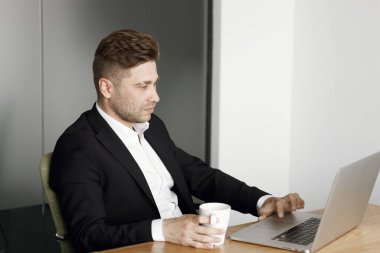 Bir adam masaya oturur ve bir fincan kahveyle çalışır. Ofiste dizüstü bilgisayarla çalışan yakışıklı bir iş adamı..