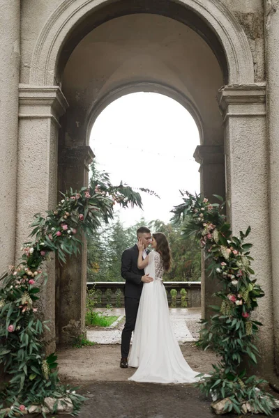 Una boda en una antigua villa, un feliz par de recién casados. Arco de boda redondo, decorado con flores blancas y vegetación, frente a la antigua arquitectura italiana. —  Fotos de Stock