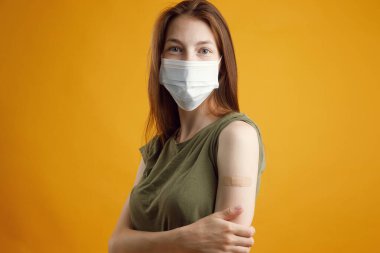 Stüdyoda sarı boş bir arka planda mutlu bir kız, aşı yapıldıktan sonra gülümsüyor ve elini gösteriyor. Coronavirus, Ncov-19, grip aşısı ve aşılama.