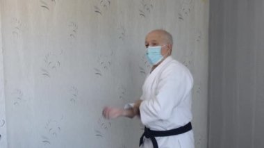 Tıbbi maske ve beyaz katategi giymiş yaşlı bir atlet blokları ve yumrukları eğitiyor.