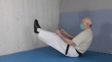 Siyah kuşak ve tıbbi bir maskeyle, tataminin üzerinde oturan yaşlı bir sporcu bacaklarını kaldırır ve aşağı iner.