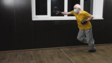 Tıbbi bir maske takan yaşlı bir sporcu kollarında siyah pedlerle yarım atlayışla bir yumruğa çarpıyor.